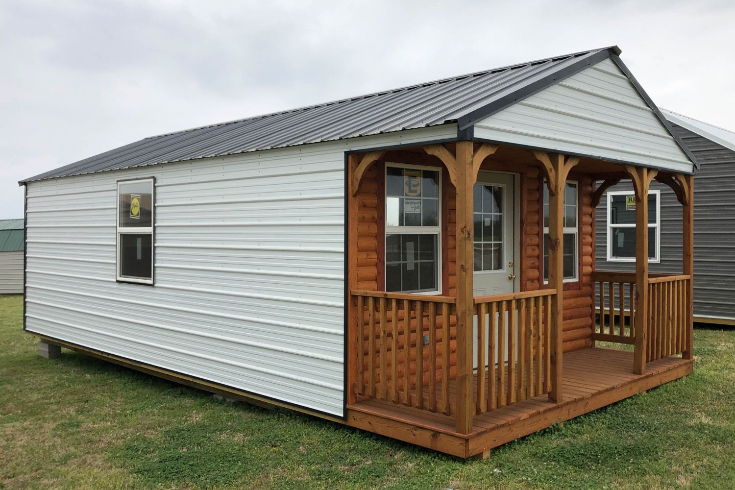 White metal cabin with a log style front porch. Cabin sites in grass on concrete block foundation